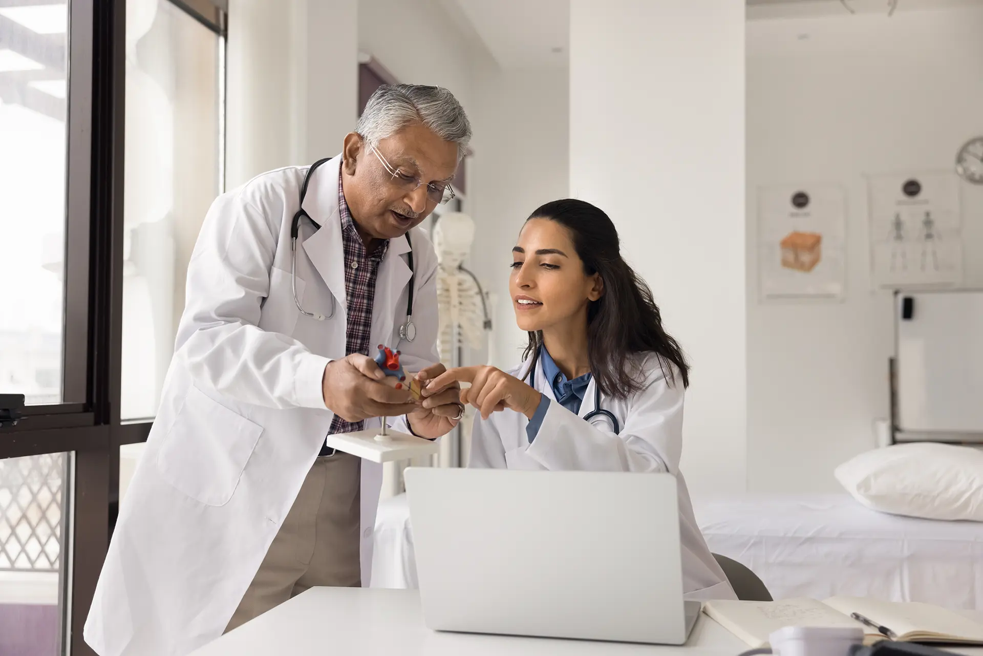 Two diverse colleagues cardiologists elderly male young female in white coats engaged in medical research study heart anatomy using model notebook collaborate discuss clinical case